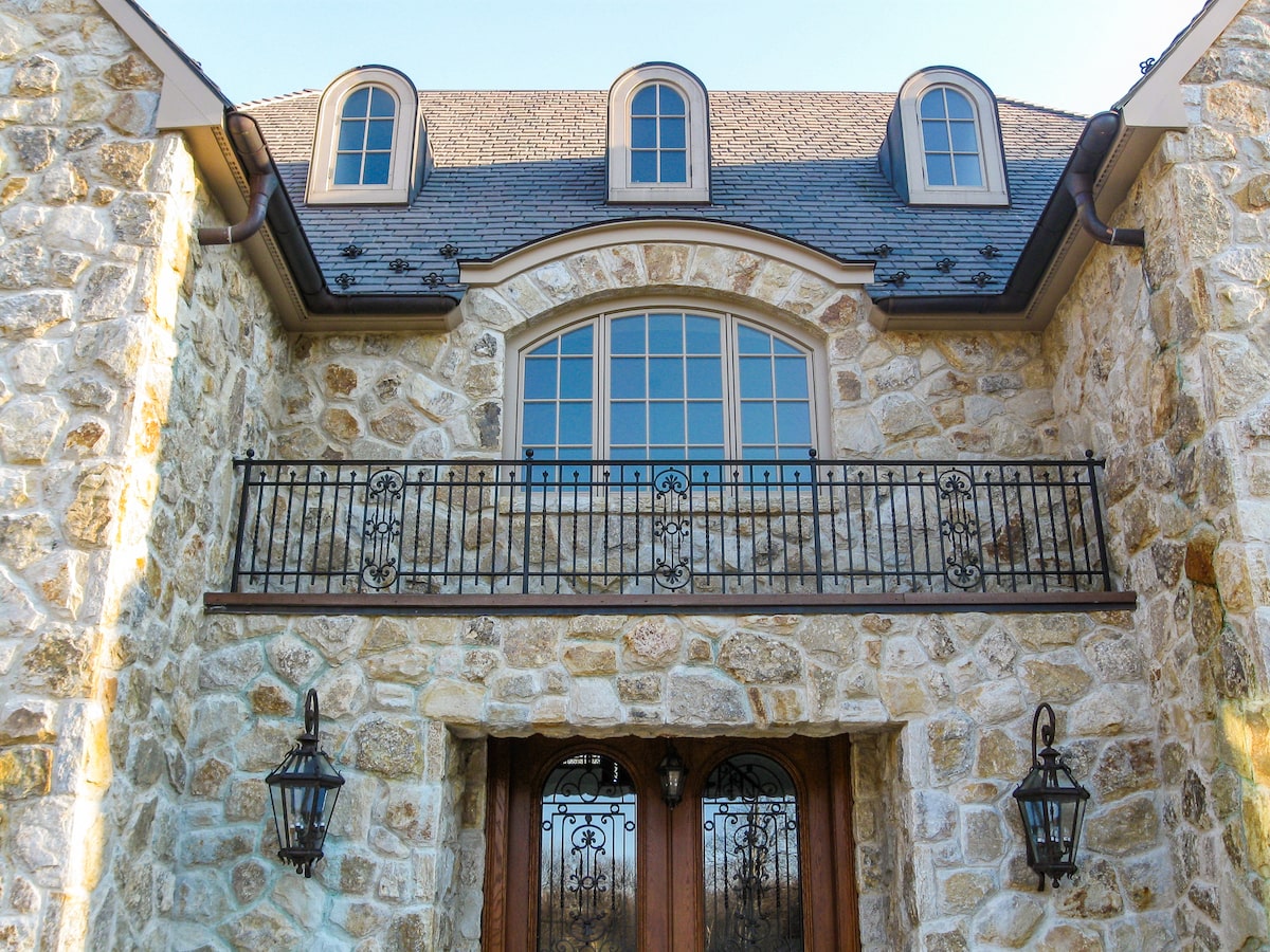 A stone house looms over the viewer, who is looking up past a double wooden door towards a false balcony below a large window. The balcony railing has vertical spindles and small decorative panels.