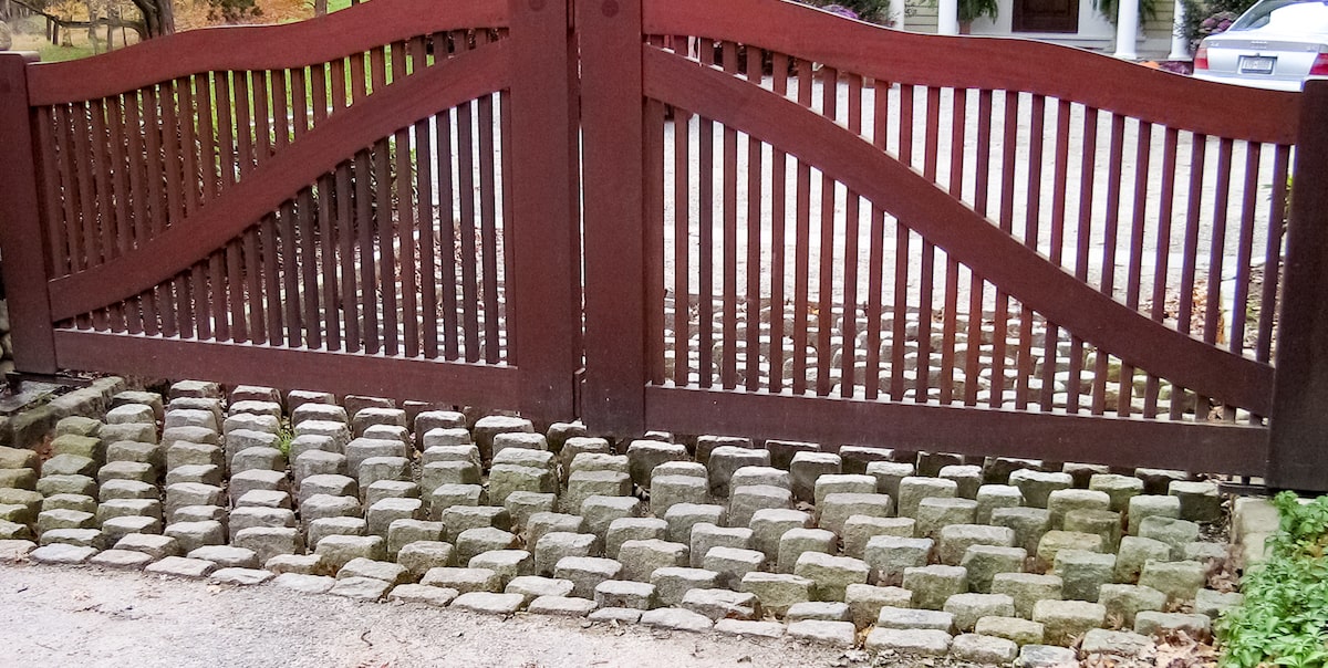 A stone cattle guard below a wooden gate. The short stones are placed vertically with gaps between them. The wooden gate has narrow vertical spindles with wide custom framing.