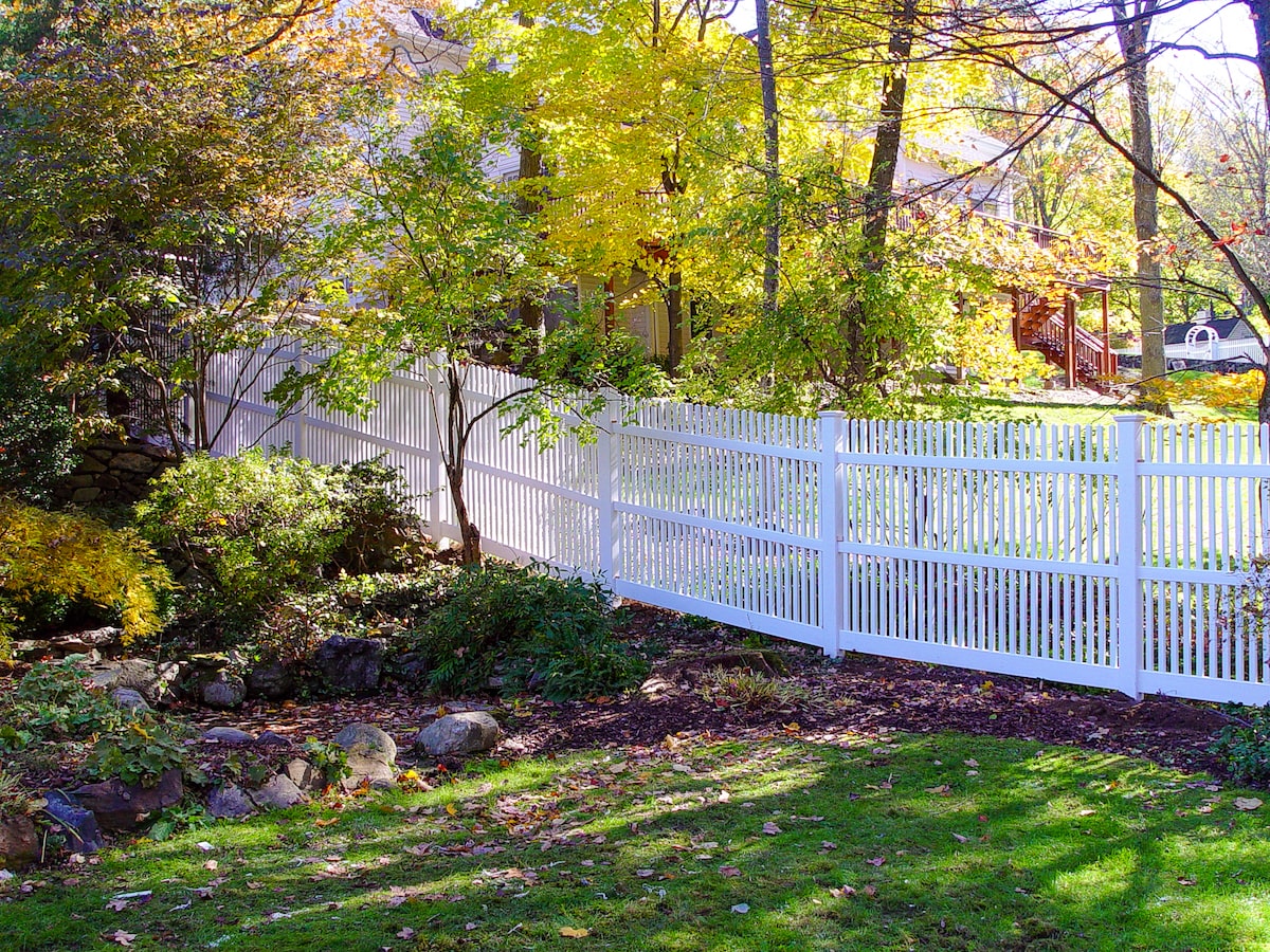 A tall white picket PVC fence surrounded by trees and bushes marks the property line between two backyards.