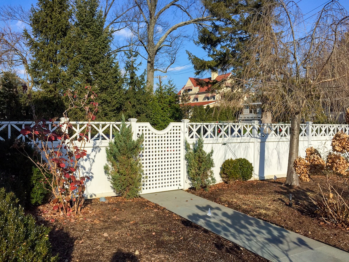 A concrete walkway leads to a white lattice gate set into a tall white privacy fence topped with decorative slats in an x pattern.