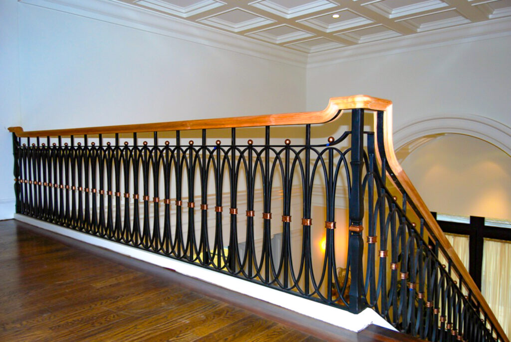 Interlocked oblong circles create a lacy pattern effect on an indoor wrought iron handrail. The viewer is positioned on a landing looking out and down the stairs.