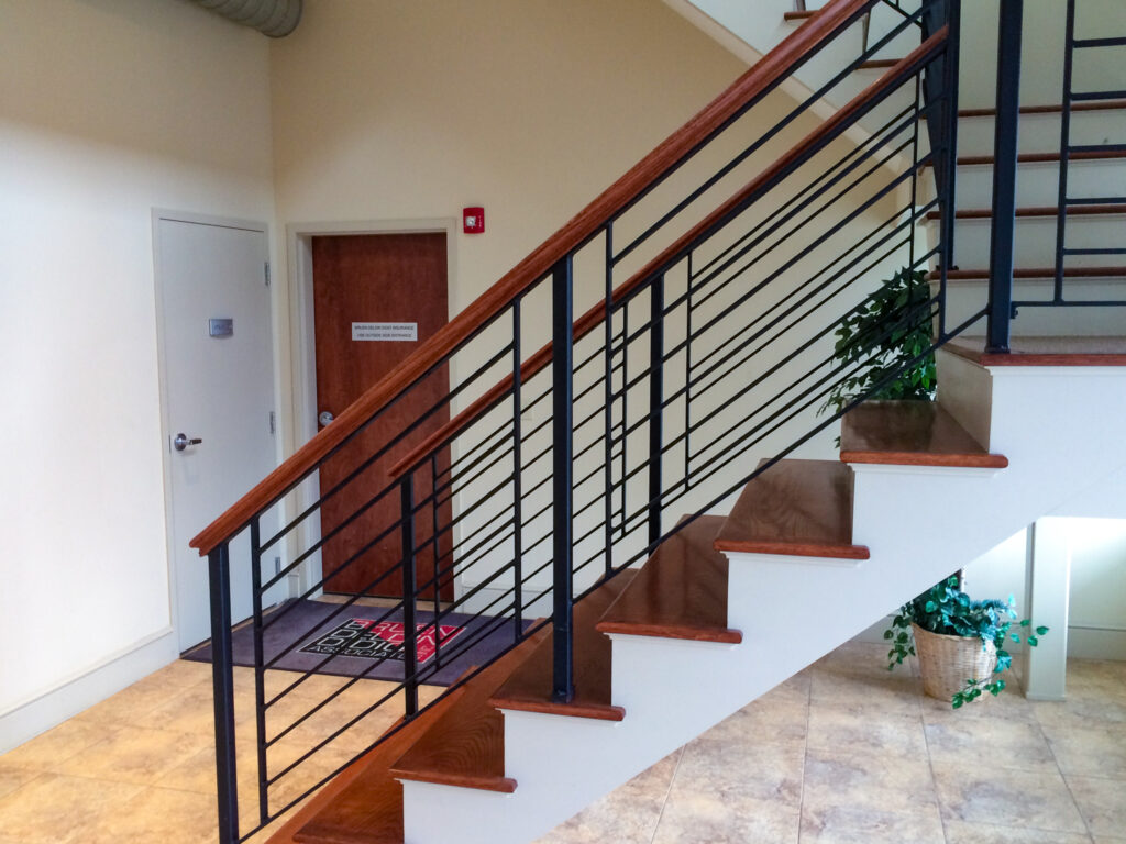 A simple black metal railing around wooden stairs in an office entryway. The handrail is wooden, and matches with the stair treads.