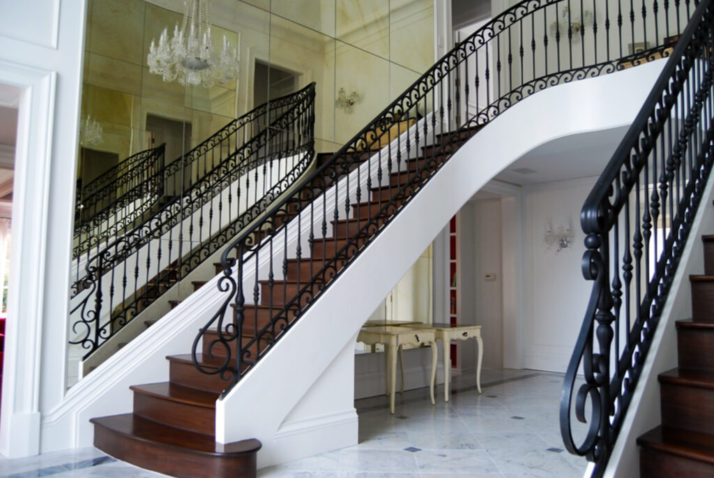 An ornate wrought iron stair railing on a double staircase creates a sweeping U shape leading up to a landing. The black of the railing contrasts with the white walls and floor as well as the dark brown wooden stairs.