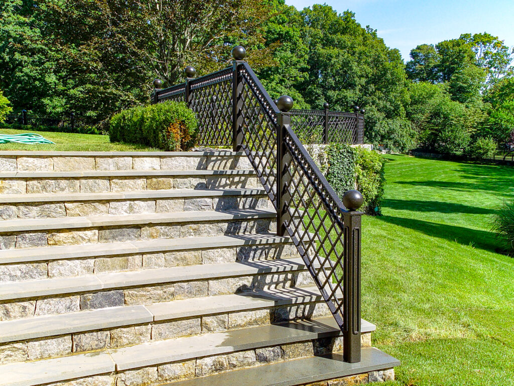 9 stone steps emerge from a sweeping green lawn. A black iron fence with diagonally-latticed panels and large spherical finials adds an architectural element to the landscape.