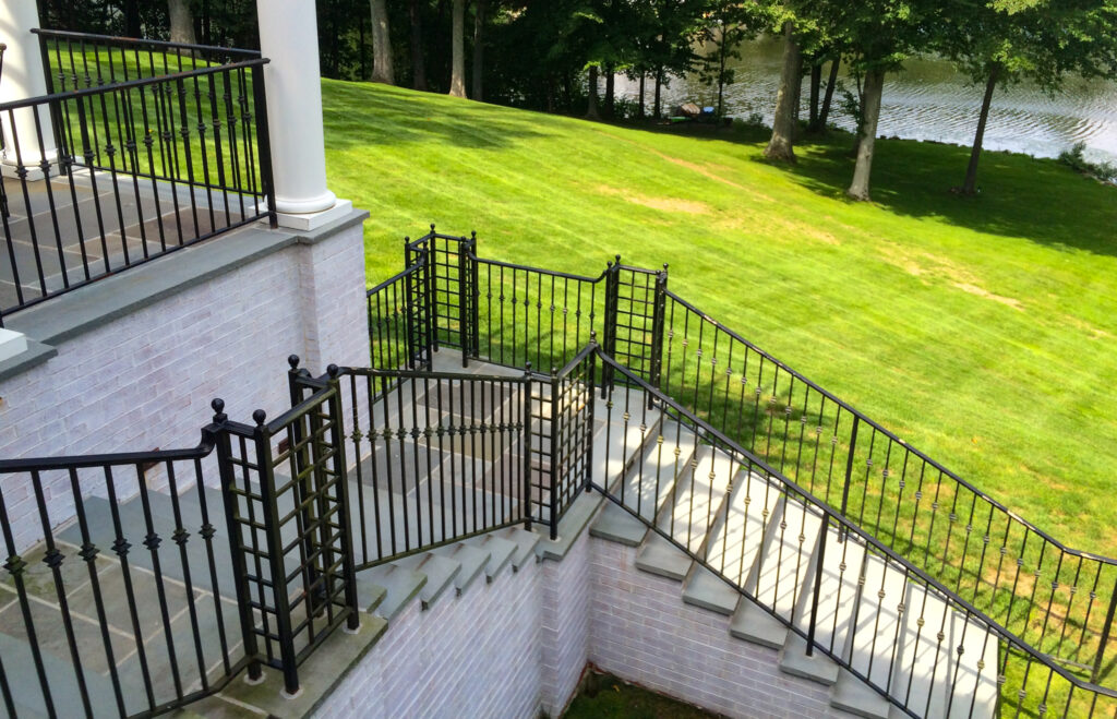 Flagstone steps lead from a high brick porch down to a patch of green grass beside trees and water. Wrought iron railings are shaped into squares at the landings, which echoes the squares of the bricks and the flagstones.