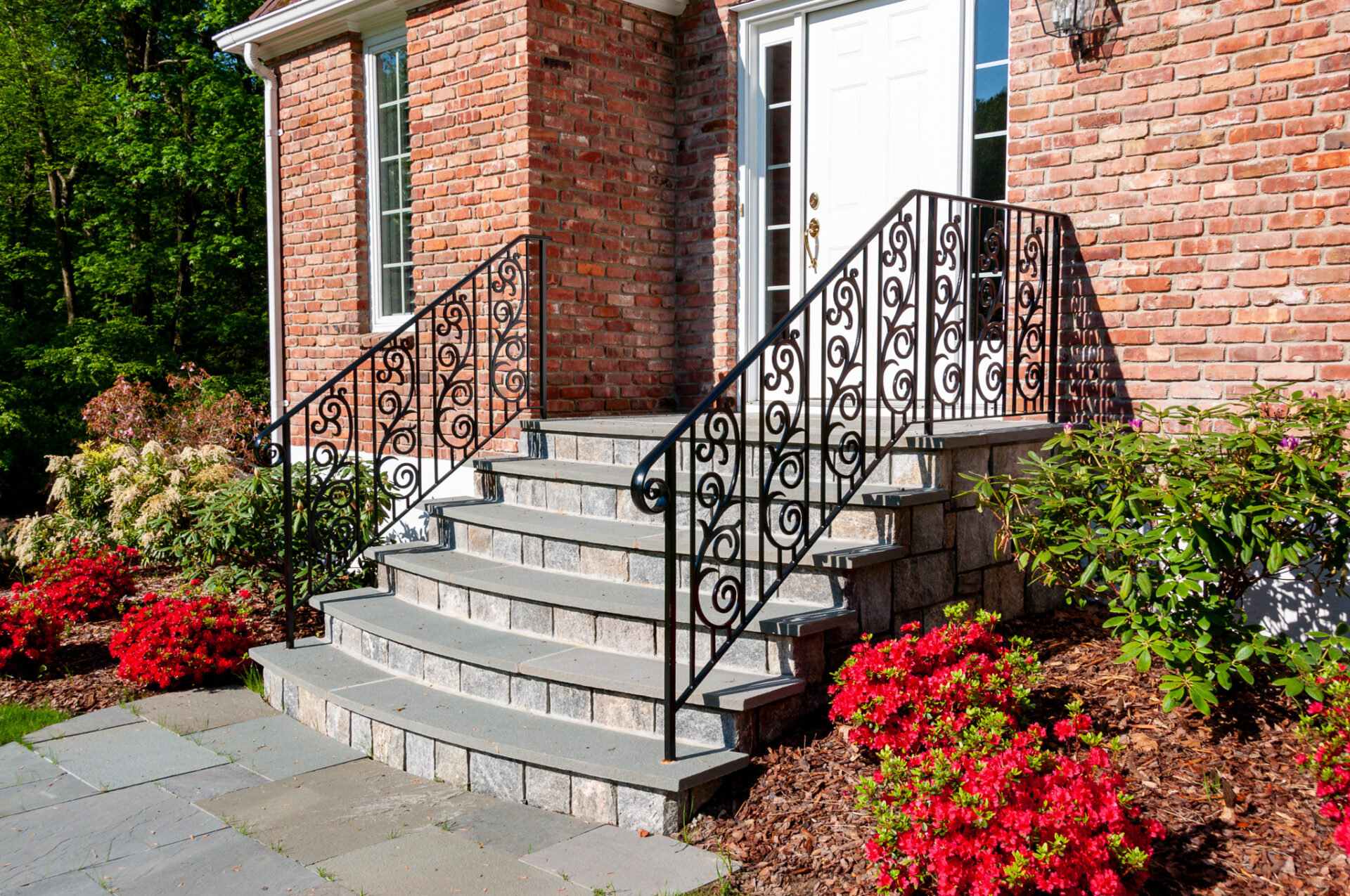 Six gray stone steps lead up to the door of a red brick house. There is an ornate black iron railing along the stairs, with the spindles shaped into vines.