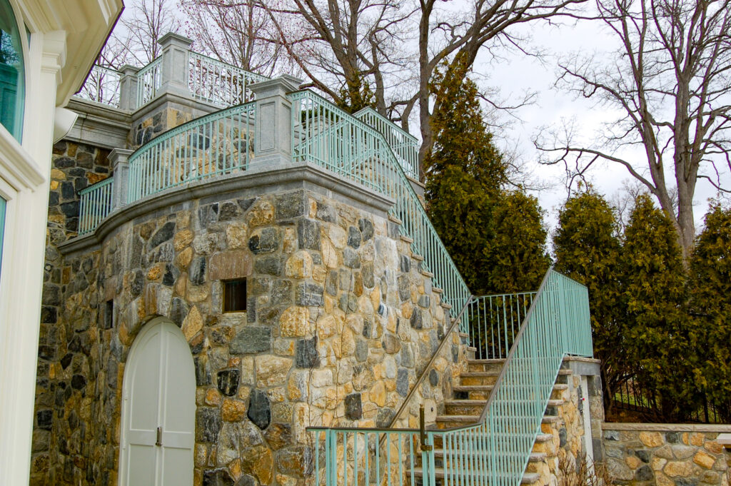 A light green metal railing protects a two-story stone staircase. The building looks like a castle or a conservatory, with arched doorways and windows.