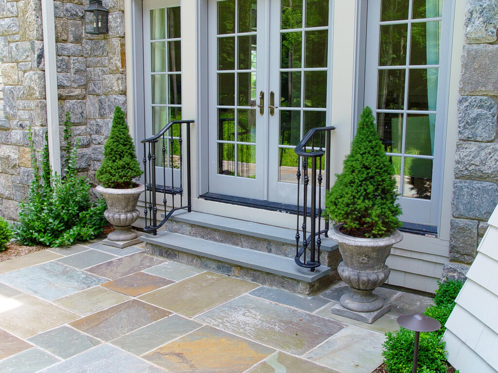 A short wrought iron railing frames three stone steps that lead up to a white French door. There are decorative planters on either side of the steps.