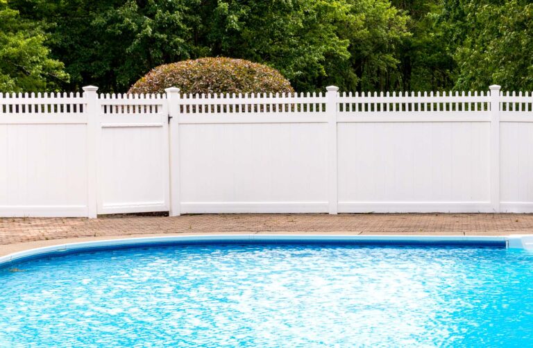 A solid white PVC fence with short pickets on top surrounds a strikingly blue pool in the foreground.