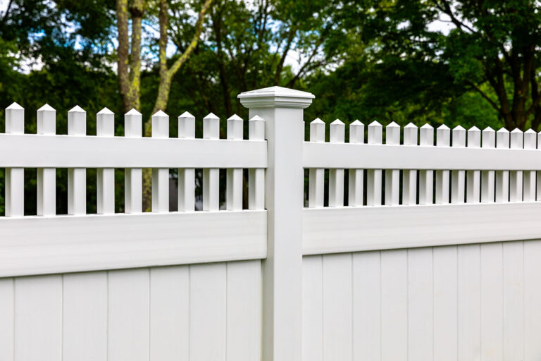 The photo shows a close up shot of a fence post cap with the short pickets on either side on top of a white solid paneled PVC fence.