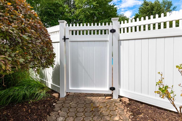 A solid panel walk gate with black metal hinges and a lock is set at an angle between the panels of a white PVC fence. A path runs up to the gate.