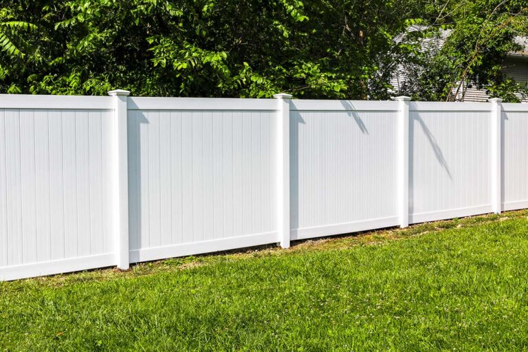 A tall white picket PVC fence surrounded by trees and bushes marks the property line between two backyards.