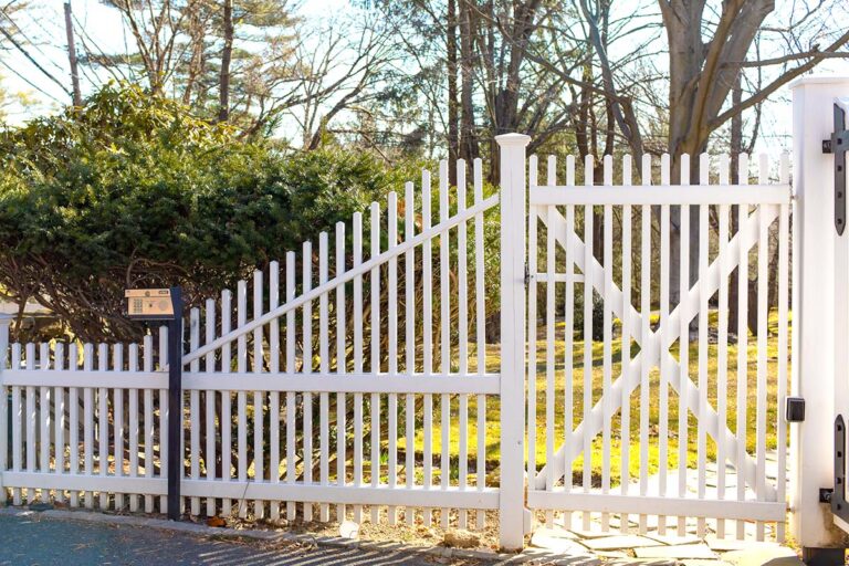 The pickets on a white PVC fence rise up quickly at an angle to a fence gate leading to a slate pathway across a lawn. There’s a call box in front of the fence and the column of a driveway gate on the far right.