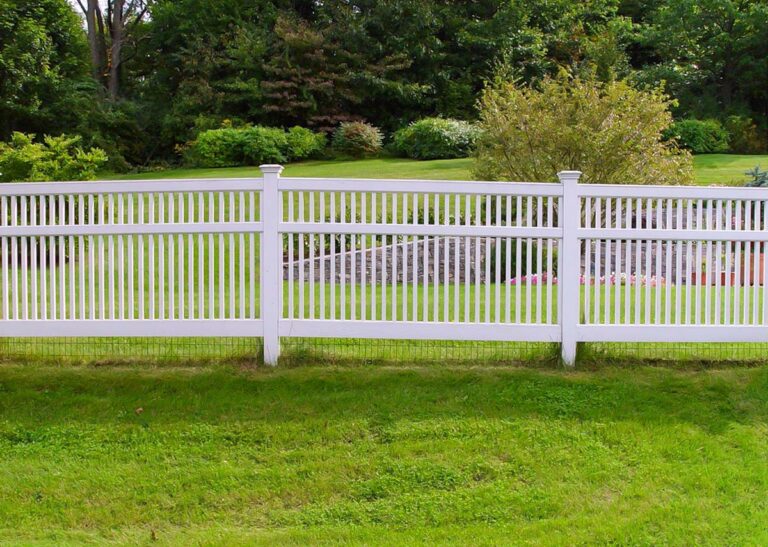 White PVC fencing extends across a lawn with a landscaped tree line in the background.